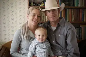 A smiling family is seated together in a cozy room. The woman has blonde hair and wears a grey top, the man wears a cowboy hat and grey shirt, and they are holding a young child who is dressed in a plaid shirt. A bookshelf with colorful books is in the background.