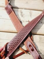 A close-up of a brown leather strap with intricate woven detailing, placed on a wooden surface. The strap has white stitching along the edges and appears to be part of a belt or bag handle, with some parts overlapping.