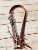 A reddish-brown leather belt with a silver buckle lies on a wooden surface. The belt features intricate patterns along its length and has leather tassels with antler tips hanging from it.