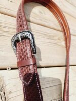 A close-up image of a brown leather belt with a decorative silver buckle. The belt features a woven pattern texture and is laid out on a wooden surface. The edges of the belt are stitched with contrasting white thread.