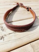 A brown leather dog collar with decorative stitching is laid on a wooden surface. The collar has a buckle and metal ring for attaching a leash. The wood planks have a rustic and weathered appearance.