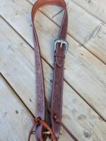 A brown leather belt with intricate carved designs and a silver ornate buckle is laid out on a wooden surface. The belt's design includes stitching along the edges and additional leather ties at one end.