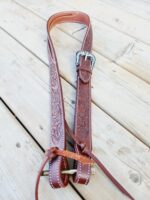 A brown leather guitar strap with intricate floral designs and a shiny silver buckle lays on a wooden floor. The strap is adjustable with leather ties at the ends.