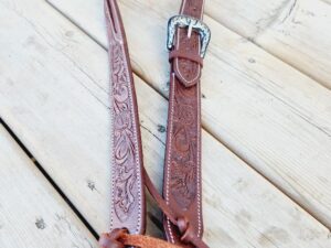 A brown leather guitar strap with intricate floral designs and a shiny silver buckle lays on a wooden floor. The strap is adjustable with leather ties at the ends.