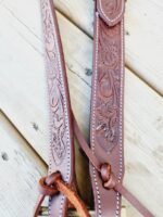 Close-up image of intricately tooled brown leather straps with floral and geometric patterns, lying on a wooden surface. The straps feature white stitching along the edges and are fastened with leather knots.