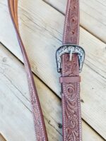 A brown leather belt with intricate embossed patterns and a decorative silver buckle featuring floral designs, resting on a wooden surface. The belt's detailed craftsmanship is highlighted by the close-up angle of the photo.