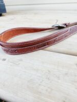 A close-up of a brown leather belt with decorative stitching and the words "RANCH BRAND" embossed on it. The belt is resting on a light wooden surface.