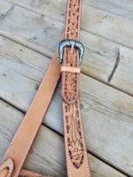 A close-up image of intricately carved leather straps with floral designs. The straps feature a silver buckle with a detailed design. The background is a wooden surface with visible grain and texture.