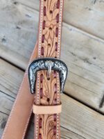 A close-up view of a leather belt with intricate floral engravings and a decorative silver buckle resting on a wooden surface. The belt features detailed craftsmanship with floral patterns and stitching along the edges.