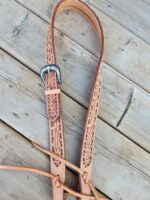 A detailed leather belt with intricate floral engravings and a silver buckle lies coiled on a wooden surface. The leather is tan with darker accents highlighting the patterns, and the belt features long, slender straps extending from it.