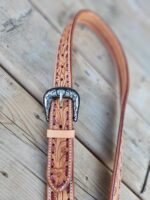 A detailed leather belt is laid out on a wooden surface. The belt features intricate floral tooling and a silver buckle with ornate designs. The belt is light brown in color, showcasing careful craftsmanship and design.