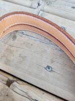 Close-up of a curved, tan leather strap with white stitching, placed on a weathered wooden surface. The image highlights the texture of the leather and the detail in the stitching. The wood surface shows visible knots and a weathered appearance.