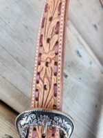 Close-up of a tooled leather belt with intricate floral patterns and a silver buckle. The leather is light brown with detailed carvings, and the buckle features ornate designs. The belt is placed on a wooden surface.