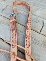 A close-up of a leather belt with intricate floral tooling, lying on a wooden surface. The belt features a detailed metal buckle and stitching along the edges, showcasing fine craftsmanship.