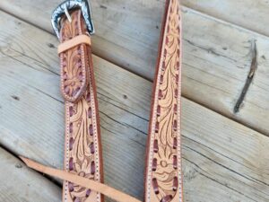 A close-up of a leather belt with intricate floral tooling, lying on a wooden surface. The belt features a detailed metal buckle and stitching along the edges, showcasing fine craftsmanship.