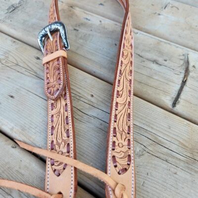 A close-up of a leather belt with intricate floral tooling, lying on a wooden surface. The belt features a detailed metal buckle and stitching along the edges, showcasing fine craftsmanship.