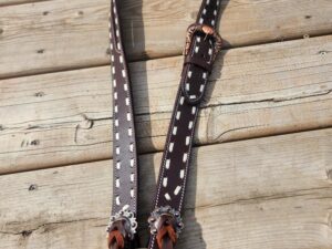 A dark brown leather belt with white stitching and a silver buckle featuring a decorative metal design is laid out on a wooden surface. The belt has two attached leather straps at the ends.