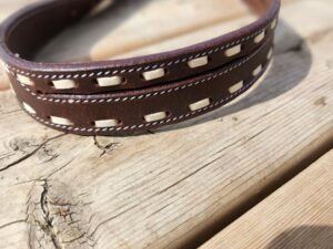 A close-up of a brown leather belt with white stitching and cut-out design, lying on a wooden surface. The belt is slightly curled, and the texture of the wooden surface is visible, with a prominent grain pattern and a crack.