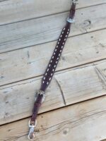 A brown leather belt with intricate silver buckles and rivets is laid out on a wooden surface. The belt features a decorative white lace design along its middle.