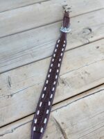 A detailed brown leather dog collar with white stitches and a silver buckle is laid out on a wooden surface. The collar features an intricate leaf design pattern along its length.