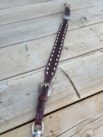 A brown leather belt with intricate white stitching and silver buckles lies on a wooden surface. The belt features detailed metal accents and a braided design in the middle section. The wood background shows a rustic, natural texture with visible knots and grain.