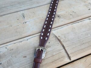 A brown leather belt with intricate white stitching and silver buckles lies on a wooden surface. The belt features detailed metal accents and a braided design in the middle section. The wood background shows a rustic, natural texture with visible knots and grain.