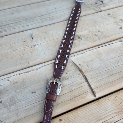 A brown leather belt with intricate white stitching and silver buckles lies on a wooden surface. The belt features detailed metal accents and a braided design in the middle section. The wood background shows a rustic, natural texture with visible knots and grain.