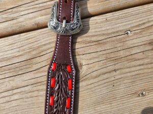 A close-up of a brown leather belt with intricate patterns and red accents. The buckle features detailed silver engravings and the belt lies on a wooden surface.