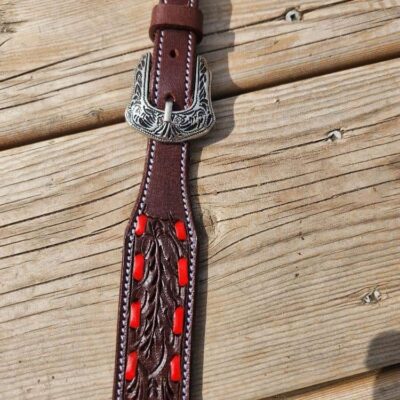 A close-up of a brown leather belt with intricate patterns and red accents. The buckle features detailed silver engravings and the belt lies on a wooden surface.