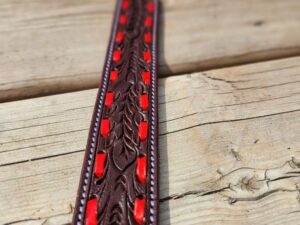 A close-up view of a brown leather belt with intricate patterns and red accents laid on a wooden surface. The background features rustic wooden planks. The focus is on the central part of the belt, showcasing its detailed design.