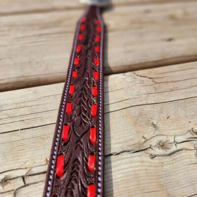 A close-up view of a brown leather belt with intricate patterns and red accents laid on a wooden surface. The background features rustic wooden planks. The focus is on the central part of the belt, showcasing its detailed design.