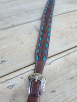 A close-up of a brown leather belt with intricate turquoise inlays and detailed stitching along the edges. The end of the belt features an ornate silver buckle with engraved floral designs, resting on a light wooden surface.