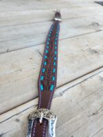 A detailed close-up of a brown leather belt with carved designs and blue accents, laid flat on a wooden surface. The belt features an ornate metal buckle with intricate patterns.