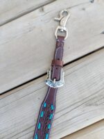 A close-up of a leather dog leash with detailed stitching and turquoise accents. The leash has a silver buckle with ornate engravings and a clasp at the end for attaching to a collar or harness. It is placed on a wooden surface.