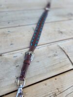 A detailed close-up of a decorative leather horse rein laid out on a wooden surface. The rein features intricate stitching, metal buckles with ornate designs, and turquoise accents woven into the leather. The background shows wooden planks with visible grains and knots.