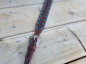 A detailed close-up of a decorative leather horse rein laid out on a wooden surface. The rein features intricate stitching, metal buckles with ornate designs, and turquoise accents woven into the leather. The background shows wooden planks with visible grains and knots.