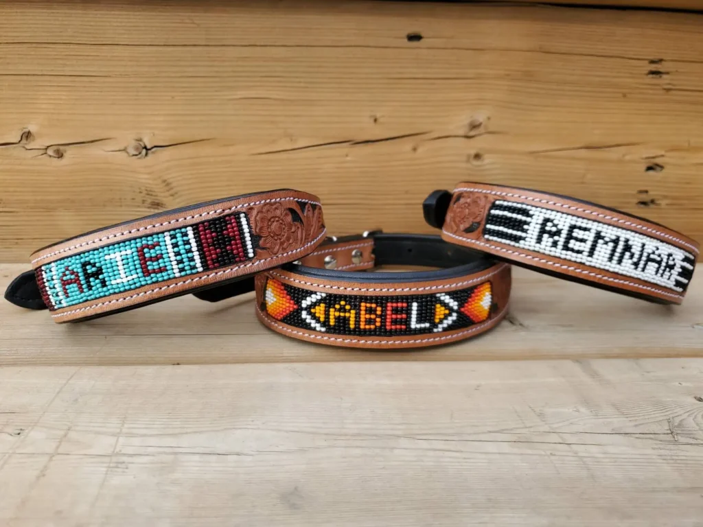 Three handmade leather dog collars with intricate beadwork are displayed on a wooden background. The collars feature custom beaded names and geometric patterns in vibrant colors, showcasing detailed craftsmanship and personal design.