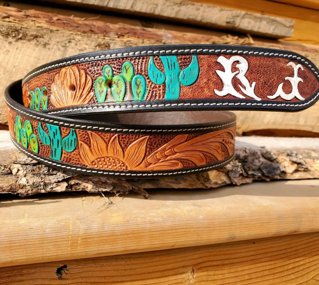 A brown leather belt with intricate embossed designs featuring a sun, cacti, and stylized patterns in black, green, blue, and white, placed on a rustic wooden surface with logs in the background.