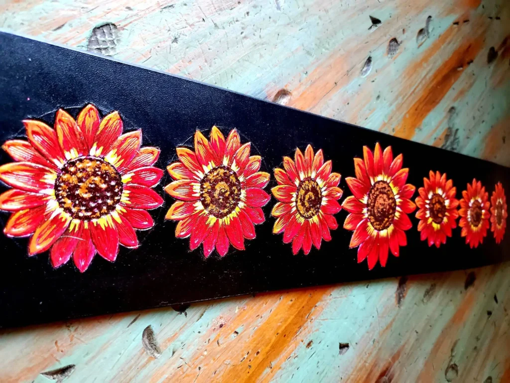 A painted strip displays a row of eight vibrant sunflowers with rich yellow, orange, and red hues against a black background. The artwork is positioned on a rustic, weathered wooden surface with light blue and orange tones.