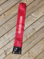 A red keychain or tag with the words "HELL BITCH" written in bold white letters is placed on a wooden surface. The tag has black fringe at one end.