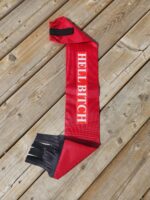 A red scarf with the words "HELL BITCH" in white text sprawled across its length lies on a wooden surface. The scarf has black fringes on one end and a black strap with Velcro on the other.