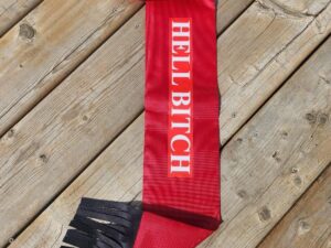 A red scarf with the words "HELL BITCH" in white text sprawled across its length lies on a wooden surface. The scarf has black fringes on one end and a black strap with Velcro on the other.