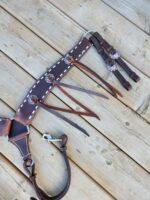 A set of brown leather equestrian tack, including bridles and reins, laid out on a wooden surface. The leather features white stitching and decorative metal conchos. Straps and buckles are visible for attaching the pieces to a horse's harness.