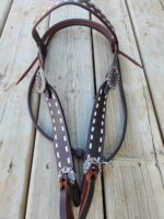 A close-up of a dark brown leather horse bridle with decorative stitching and metal buckles. The bridle is lying on a wooden surface, showcasing its craftsmanship and details.