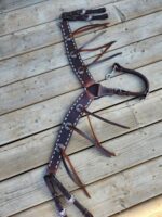 A brown leather horse breast collar with intricate white stitching and silver conchos lies spread out on a wooden surface. Several straps extend from the main piece, each with detailed craftsmanship, showcasing an ornate design intended for equestrian use.