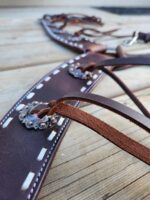 Close-up of a brown leather bridle with intricate stitching and decorative metal conchos, laying on a wooden surface. The bridle features detailed craftsmanship, including lacing and metal accents, giving it a rustic and elegant appearance.