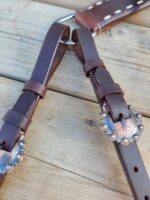 Close-up of leather saddle straps on a wooden surface, featuring decorative metal conchos with intricate designs. The dark brown leather is stitched with white thread, and the conchos add an aesthetic touch to the functional equestrian gear.