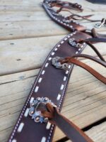 Close-up of a leather harness strap lying on a wooden surface. The strap has decorative metal conchos and white stitching. Thin leather cords are threaded through holes along the edges, adding to the intricate design.