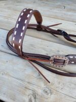 A close-up image of a brown leather horse bridle with white stitching and decorative embellishments lies on a wooden surface. The bridle includes intricate details and metal hardware.