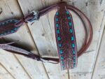 A close-up of an intricately designed leather belt with turquoise inlays and white stitching. The belt, featuring a silver buckle with engraved patterns, is laid out on a wooden surface. The detailed craftsmanship is highlighted with floral carvings on the leather.